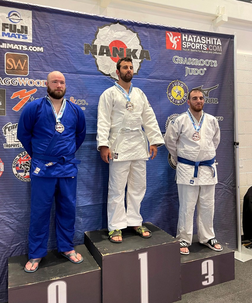 Jacob Markarian stands on the first-place podium at a judo competition, wearing a white judo gi and medal alongside two fellow competitors.